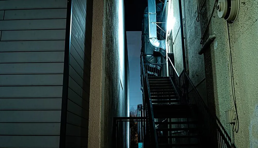 The image shows a narrow dimly lit alleyway with a metal staircase between two buildings at night