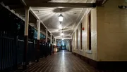 The image shows an empty, well-lit corridor with a series of hanging lanterns, lined by pillars on one side and windows on the other, evoking a serene and historical atmosphere.