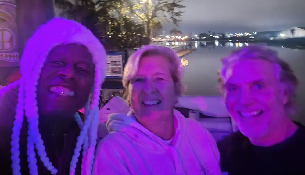 Three people are smiling for a selfie at night with purple lighting and a waterfront in the background
