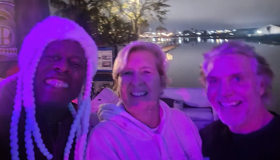 Three people are smiling for a selfie at night with purple lighting and a waterfront in the background.
