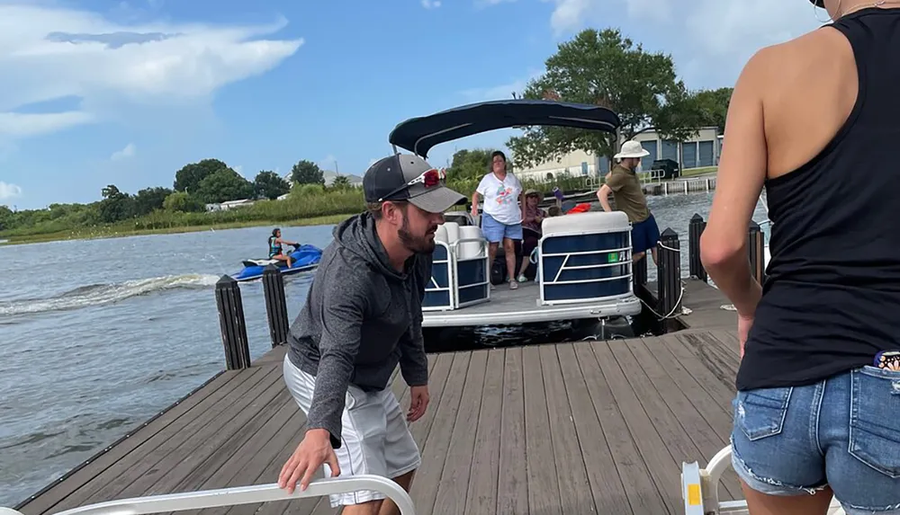 People are engaged in various activities on a sunny dock with some preparing a pontoon boat for a ride others observing and a person jet skiing in the background