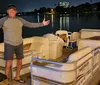 A man is standing on a dock at night beside a pontoon boat smiling and gesturing with his arms open wide
