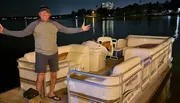 A man is standing on a dock at night beside a pontoon boat, smiling and gesturing with his arms open wide.