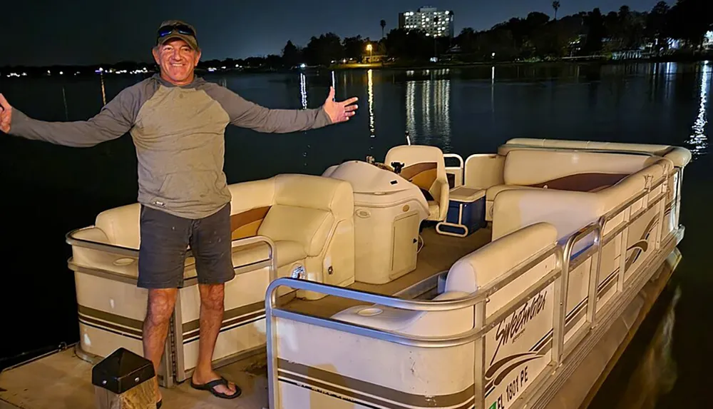 A man is standing on a dock at night beside a pontoon boat smiling and gesturing with his arms open wide