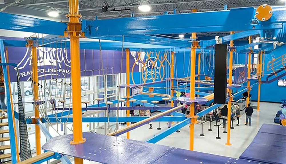 The image shows an indoor trampoline park with various climbing structures and activity zones in vibrant blue and orange colors