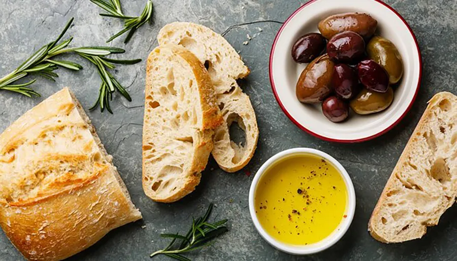 The image shows a simple and appetizing arrangement of sliced bread, a bowl of mixed olives, olive oil with pepper, and fresh rosemary on a textured surface.