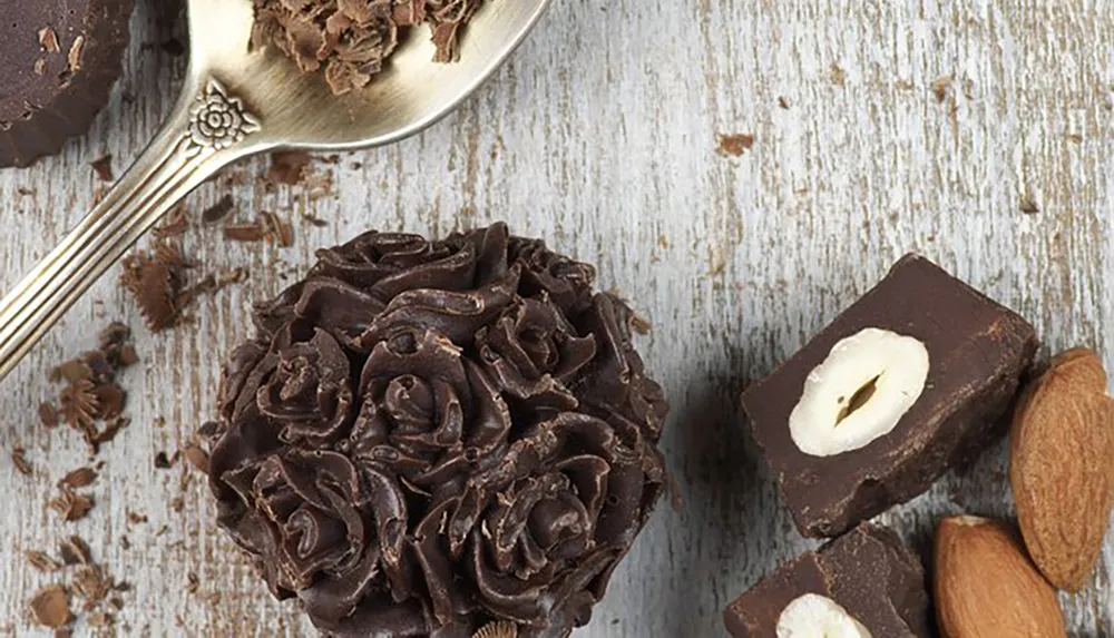 The image shows an assortment of chocolates including chocolate shavings on a silver spoon and a chocolate truffle decorated with curled chocolate shavings on a rustic wooden background