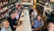 A group of people is smiling and posing for a photo at a long table in a cozy wine shop or bar.
