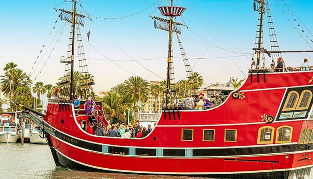 A red pirate-themed tour boat is docked at a harbor bustling with passengers on board and visitors around in a tropical setting with palm trees in the background