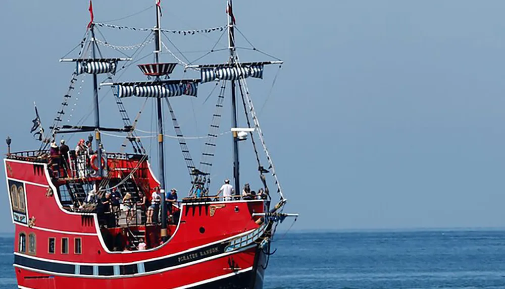 The image shows a vibrant red pirate-themed tour boat with passengers on deck sailing on a calm blue sea under a clear sky