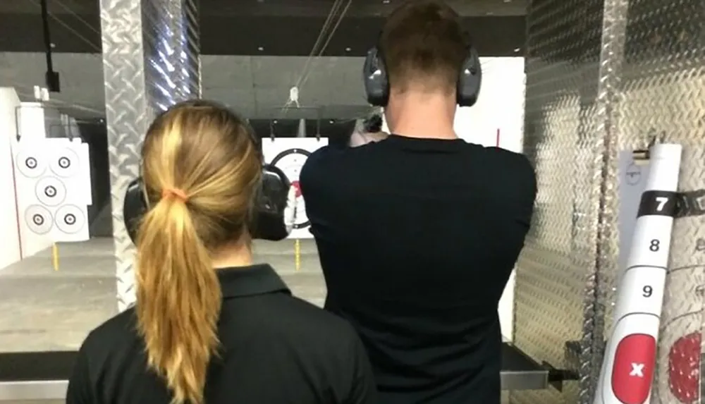 Two people are wearing protective earmuffs at an indoor shooting range observing targets downrange