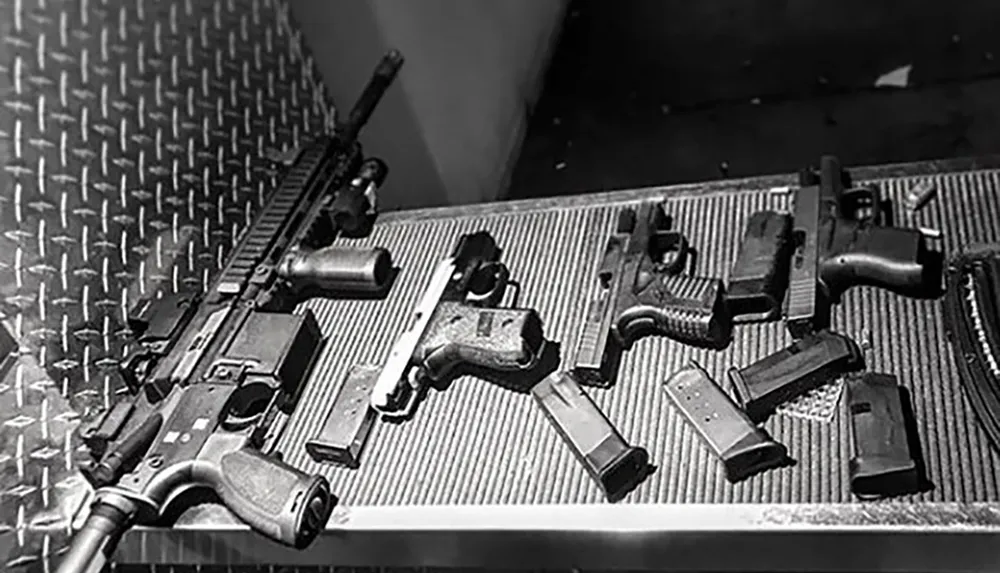 The image shows an assortment of firearms and magazines laid out on a textured surface captured in black and white