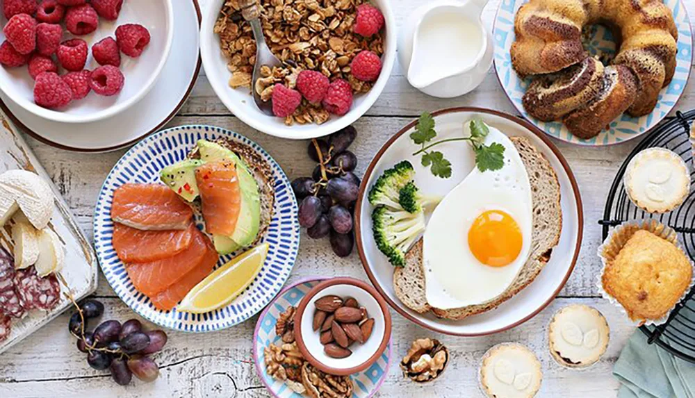 The image displays a variety of breakfast foods including fresh fruit granola with raspberries smoked salmon with avocado a sunny-side-up egg on toast with greens pastries nuts and a jug of milk all arranged on a wooden surface