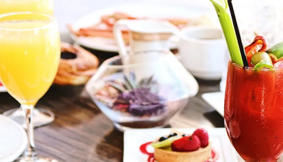 The image features a brunch setting with a bright mimosa, a Bloody Mary garnished with vegetables, and a variety of food including pastries and a fruit tart.