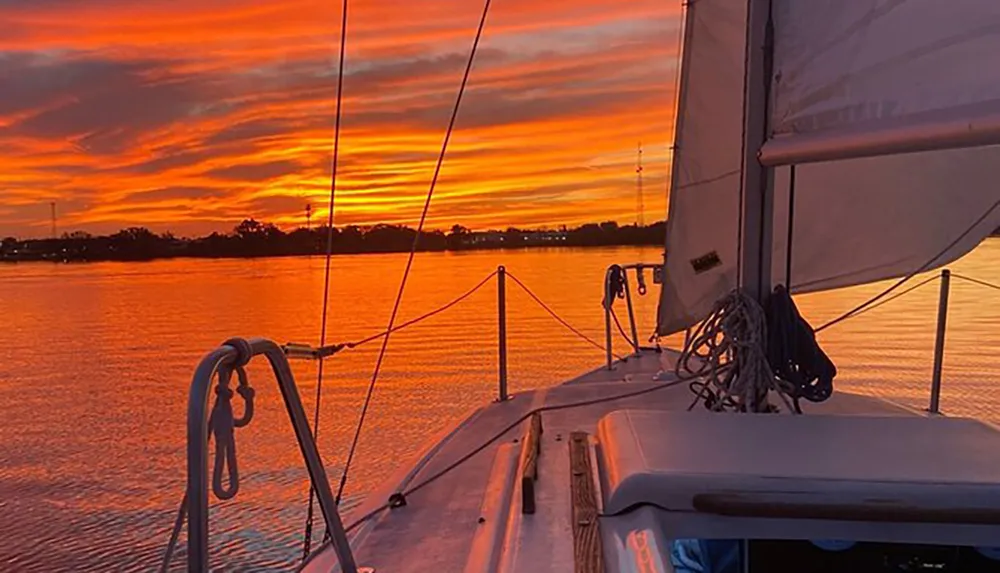 A sailboat is seen at dusk with sails partially unfurled against a stunning backdrop of a vividly colored sunset sky reflecting on calm waters