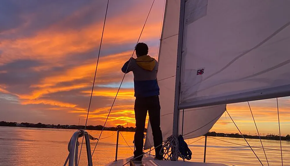 A person is standing on a sailboat holding the rigging while enjoying a stunning sunset over the water