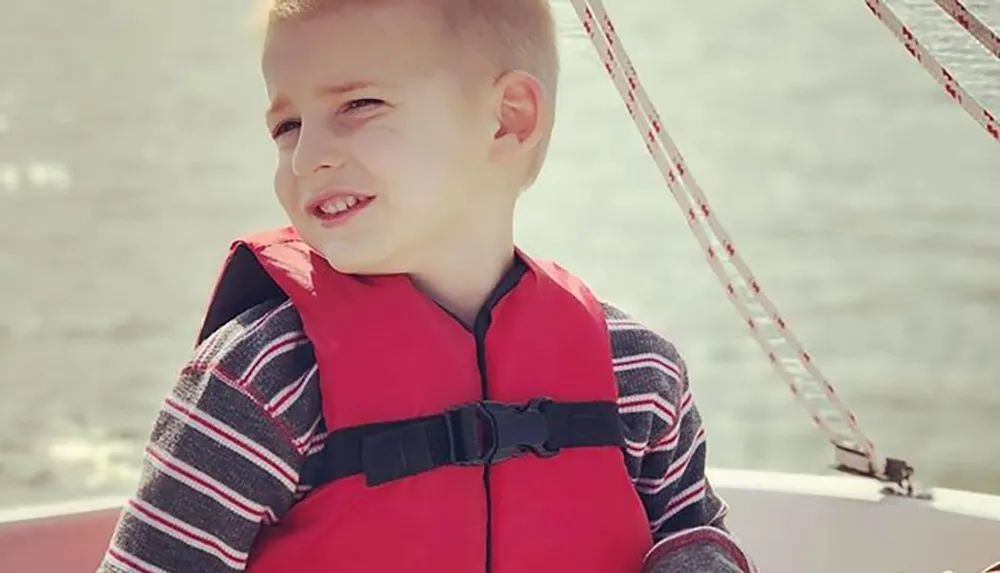A young child is wearing a red life jacket and squinting in the sunlight on a boat