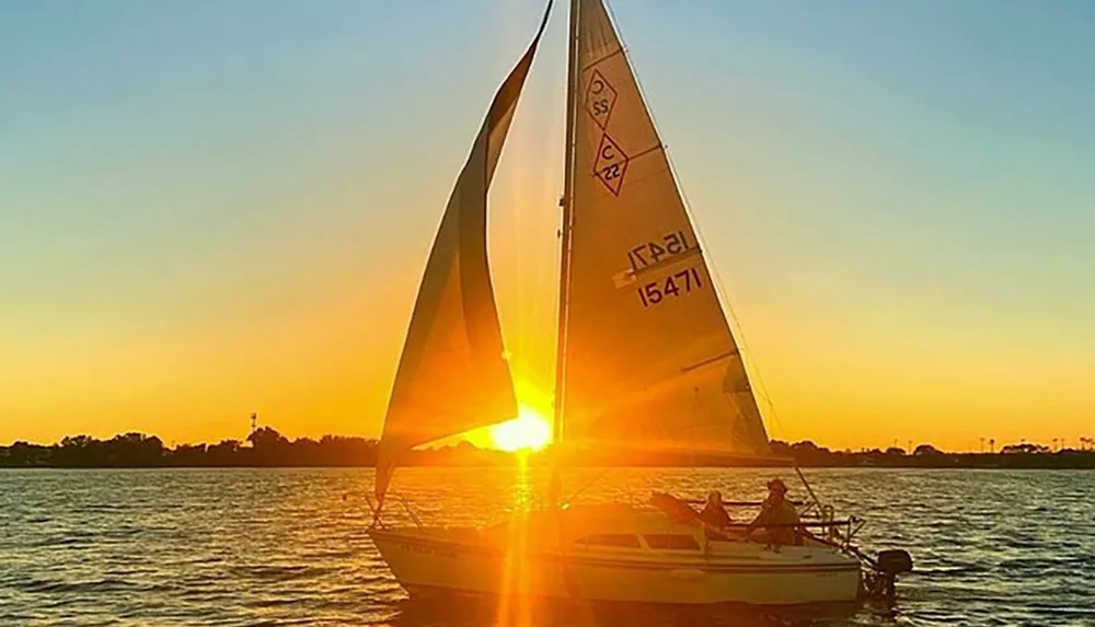 A sailboat glides on the water against a beautiful sunset backdrop with the suns rays creating a striking silhouette of the sails