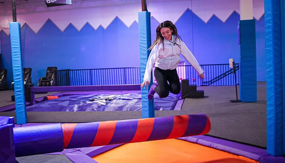 A person is joyfully jumping on a colorful indoor trampoline in a play area adorned with vibrant soft padding and walls painted with a mountain silhouette