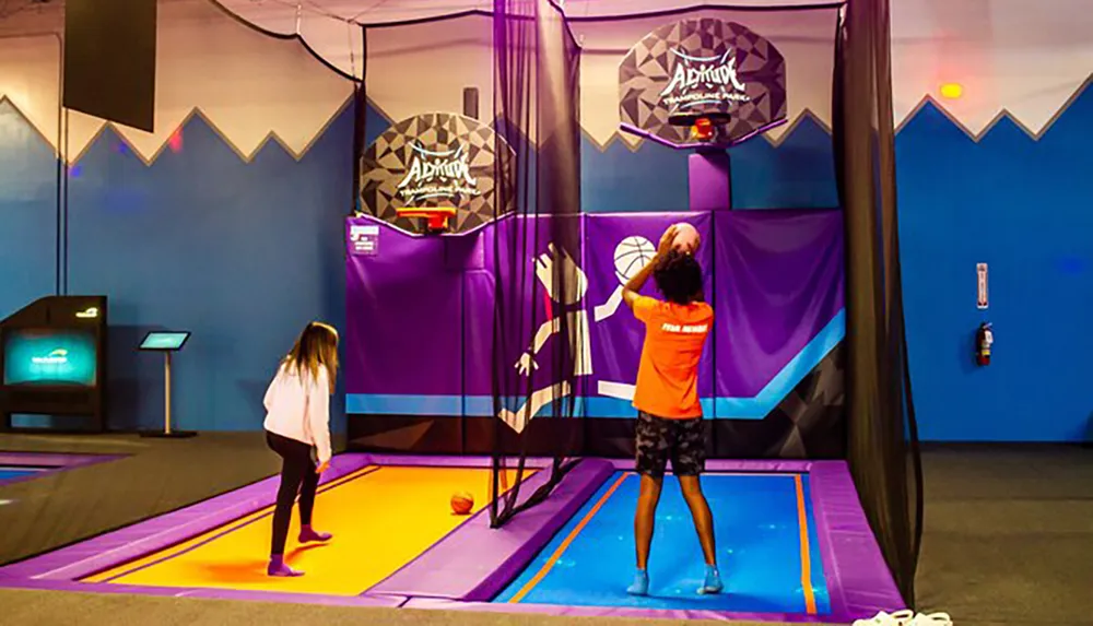 A person in an orange staff t-shirt is playing basketball on a trampoline while another person watches at an indoor trampoline park