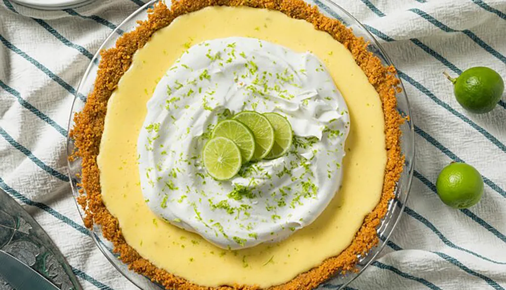 The image shows a freshly prepared key lime pie with whipped cream and lime slices on top resting on a blue and white striped cloth with whole limes nearby