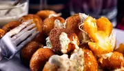 The image shows a plate of golden brown, crispy fried snacks, possibly cheese fritters, with one of them split open to reveal a gooey, melted center, served with a side of sauce or dip.