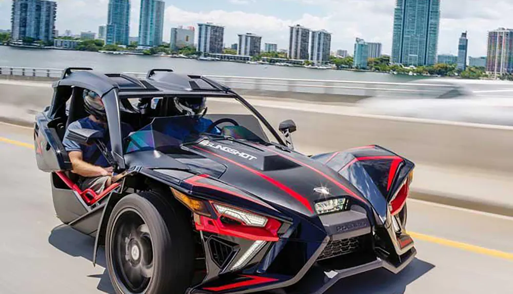 A person is driving a three-wheeled Polaris Slingshot on a bridge with a city skyline in the background