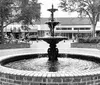 A man wearing a cowboy hat and a Sanford Tours  Experiences t-shirt is speaking while gesturing with his hands standing in an outdoor plaza with a fountain and string lights in the blurred background