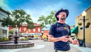 A man wearing a cowboy hat and a Sanford Tours & Experiences t-shirt is speaking while gesturing with his hands, standing in an outdoor plaza with a fountain and string lights in the blurred background.