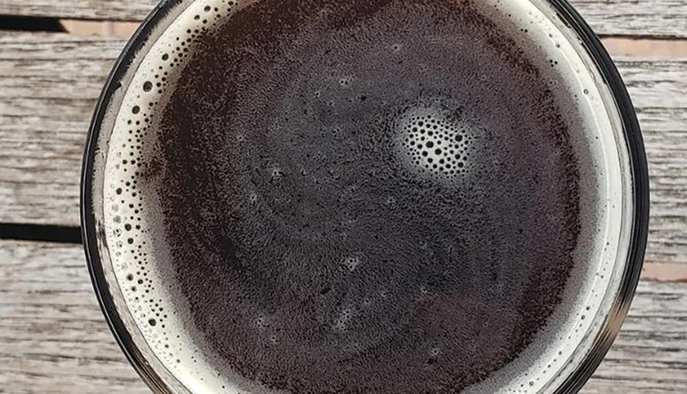 The image shows a top-down view of a dark beverage with a foamy head possibly a stout or porter beer in a glass placed on a wooden surface
