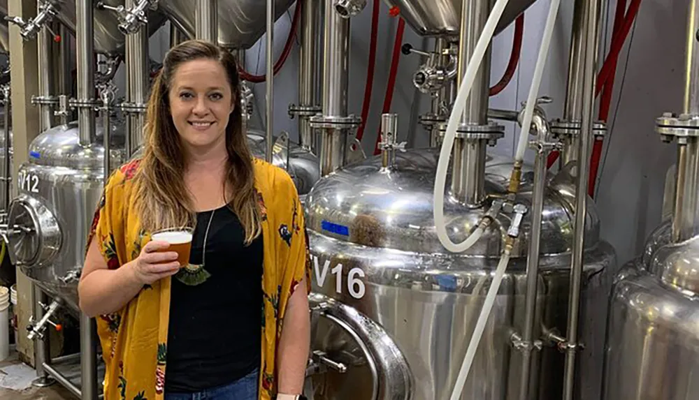 A woman is posing with a glass of beer in front of stainless steel brewing tanks in a brewery
