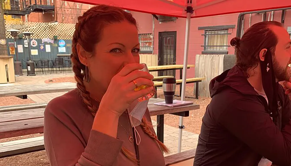 A woman with her hair in a braid is covering her nose and mouth with a napkin while holding a cup sitting at an outdoor seating area with red umbrellas and another person visible in profile