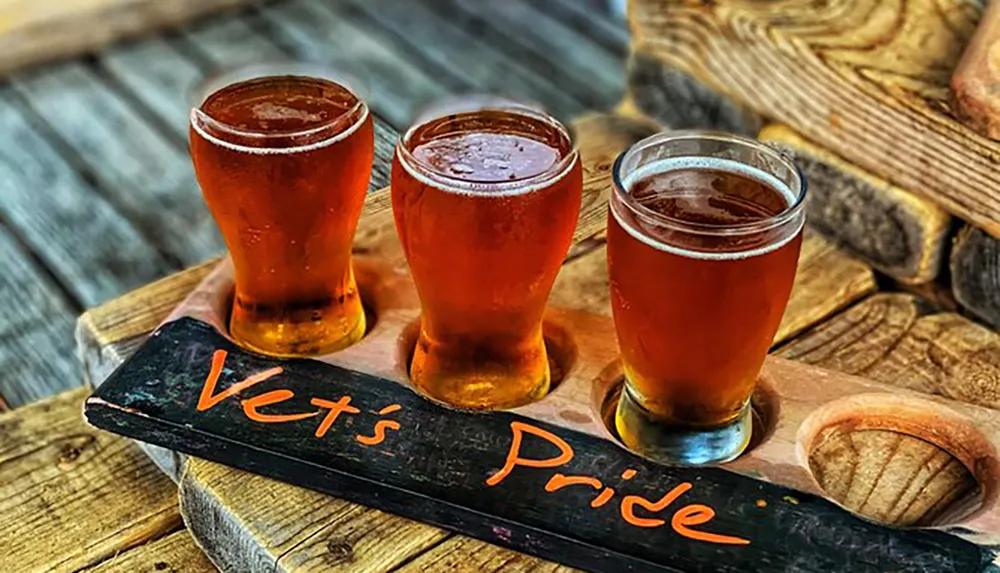 A flight of amber-colored beers is presented on a wooden paddle with the words Vets Pride written on it