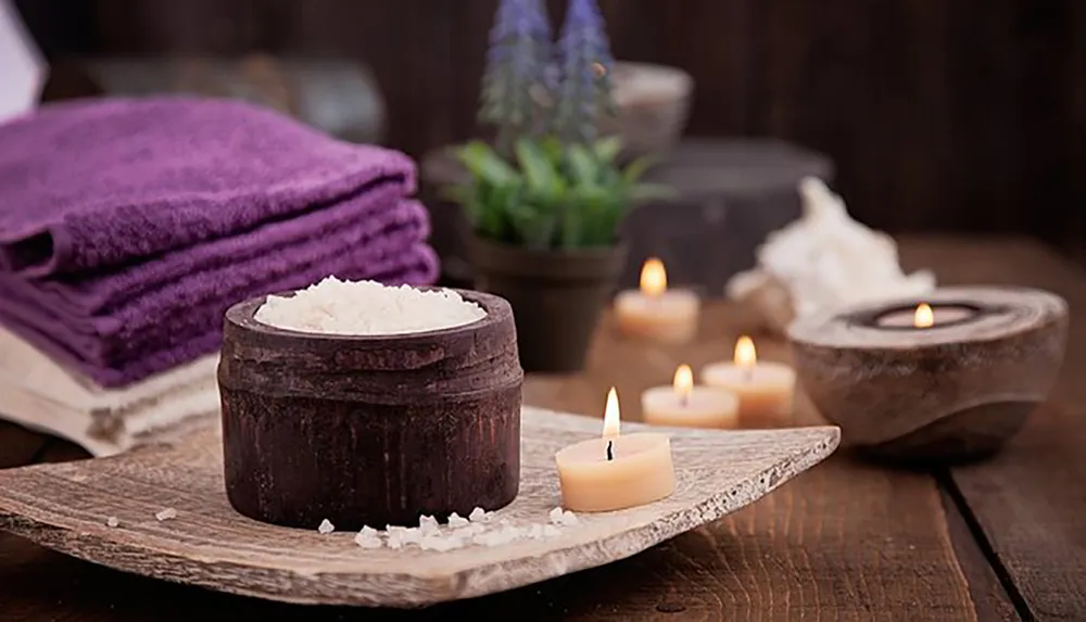 The image depicts a tranquil spa setting with a bowl of salt lit candles a stack of purple towels and a potted plant creating an atmosphere of relaxation and self-care