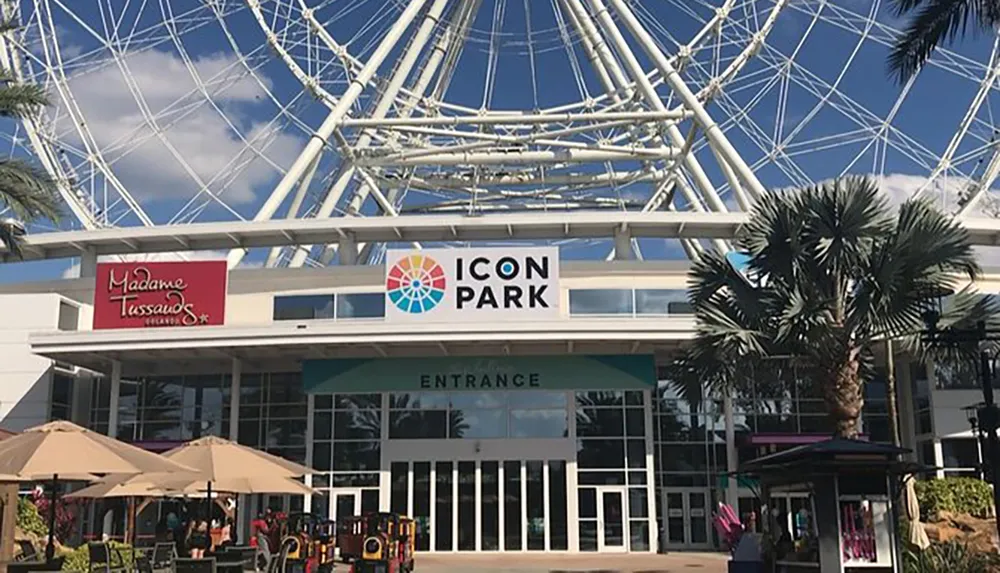The image shows the entrance to ICON Park featuring the Madame Tussauds sign with a large Ferris wheel structure visible in the background and a clear blue sky above