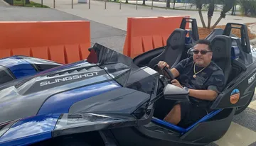 A person is seated in a parked Polaris Slingshot three-wheeled vehicle, smiling and holding the steering wheel.