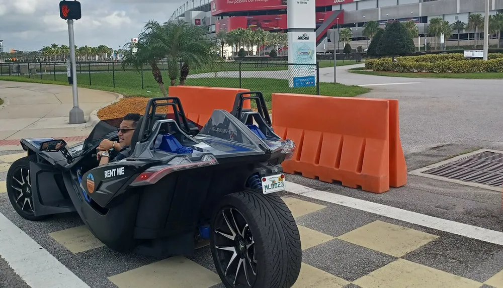 A person sits in a parked open-topped three-wheeled rental vehicle with prominent RENT ME signage at a pedestrian crossing by the Daytona International Speedway