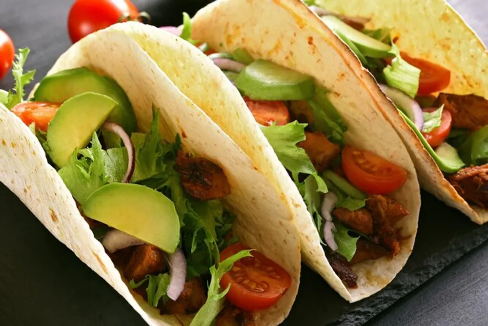 The image depicts three delicious-looking tacos filled with grilled meat slices of avocado cherry tomatoes lettuce and red onion all served on a dark slate tray