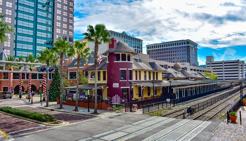The image shows a vibrant urban scene with a historic-looking train station building adorned with Christmas decorations flanked by modern high-rise buildings and palms under a partly cloudy sky