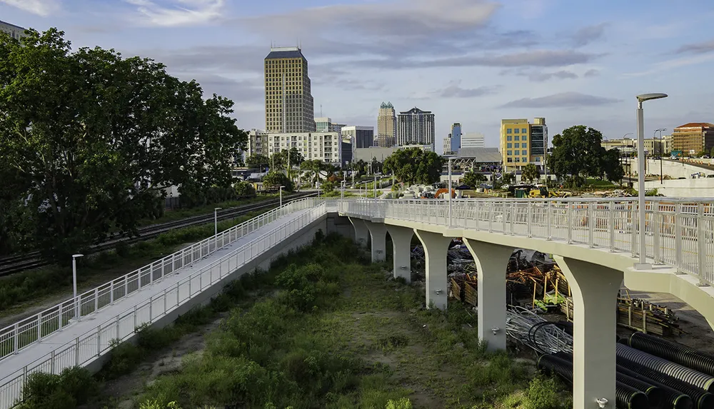 A pedestrian bridge spans a green area leading towards a urban skyline with various buildings under a cloudy sky