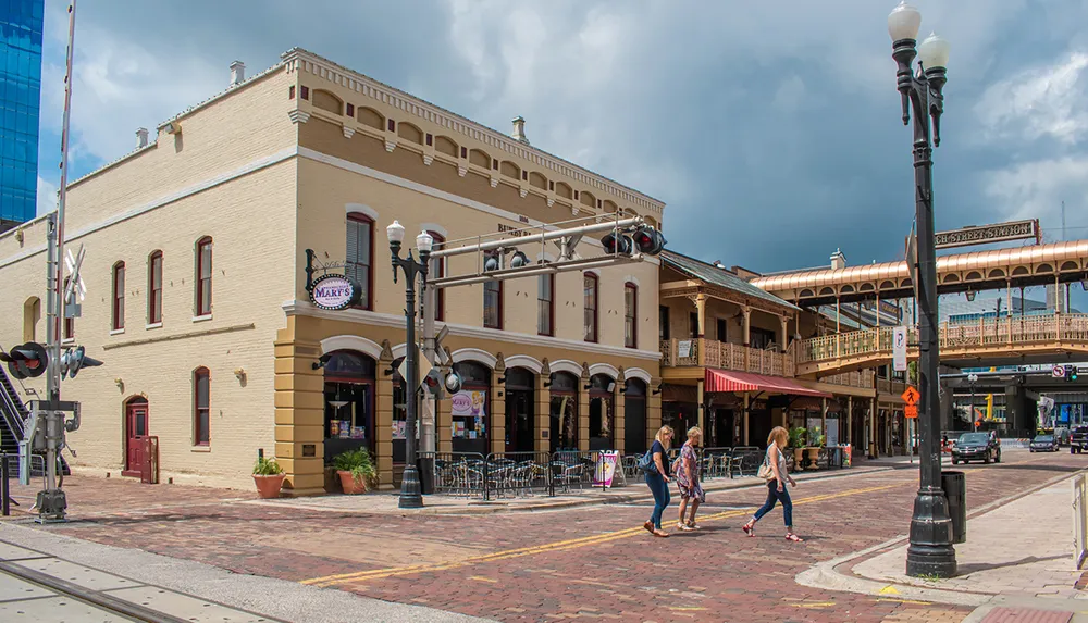 The image shows a street corner in a historic district with pedestrians crossing vintage architecture a clear sky above and a visible railway track and crossing signals in the foreground