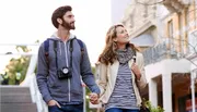 A smiling couple, with the man carrying a camera, holds hands while walking and gazing around in an urban setting during what appears to be dusk.