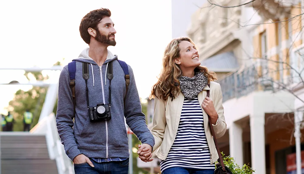 A smiling couple with the man carrying a camera holds hands while walking and gazing around in an urban setting during what appears to be dusk