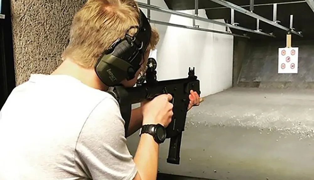 A person wearing hearing protection is aiming a rifle at a target in a shooting range