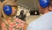 A woman is aiming a rifle at a shooting range under the supervision of an instructor.