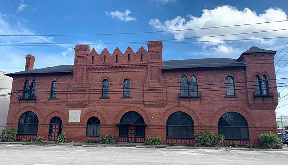 The image shows a two-story red brick building with distinctive arched windows battlements and a bell tower feature set against a blue sky with clouds