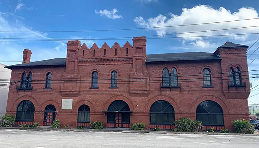 The image shows a two-story red brick building with distinctive arched windows, battlements, and a bell tower feature, set against a blue sky with clouds.