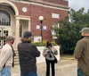 A group of people is standing outside a building listening to a woman who appears to be giving a tour or presentation