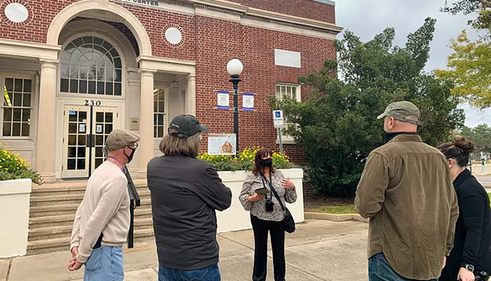 A group of people is standing outside a building listening to a woman who appears to be giving a tour or presentation