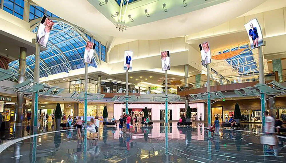 The image shows a spacious and modern shopping mall interior with a glass ceiling shiny floors shoppers and large digital screens displaying advertisements